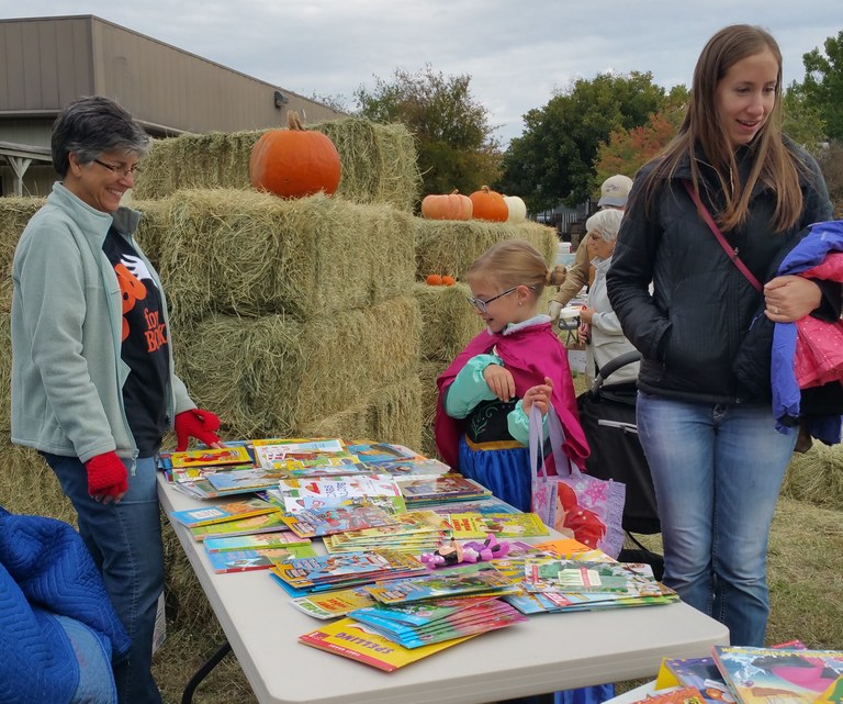 Children in Halloween costumes with their parents are invited to pick a free book from a table with a librarian. 